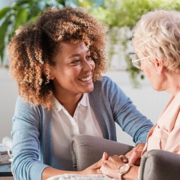 Female home caregiver talking with senior woman, sitting in living room and listening to her carefully.